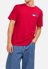 Rotes Baumwoll-T-Shirt mit kurzen Ärmeln, Rundhalsausschnitt und einem kleinen blau-weißen Logo auf der linken Brust; getragen mit blauen Jeansshorts.