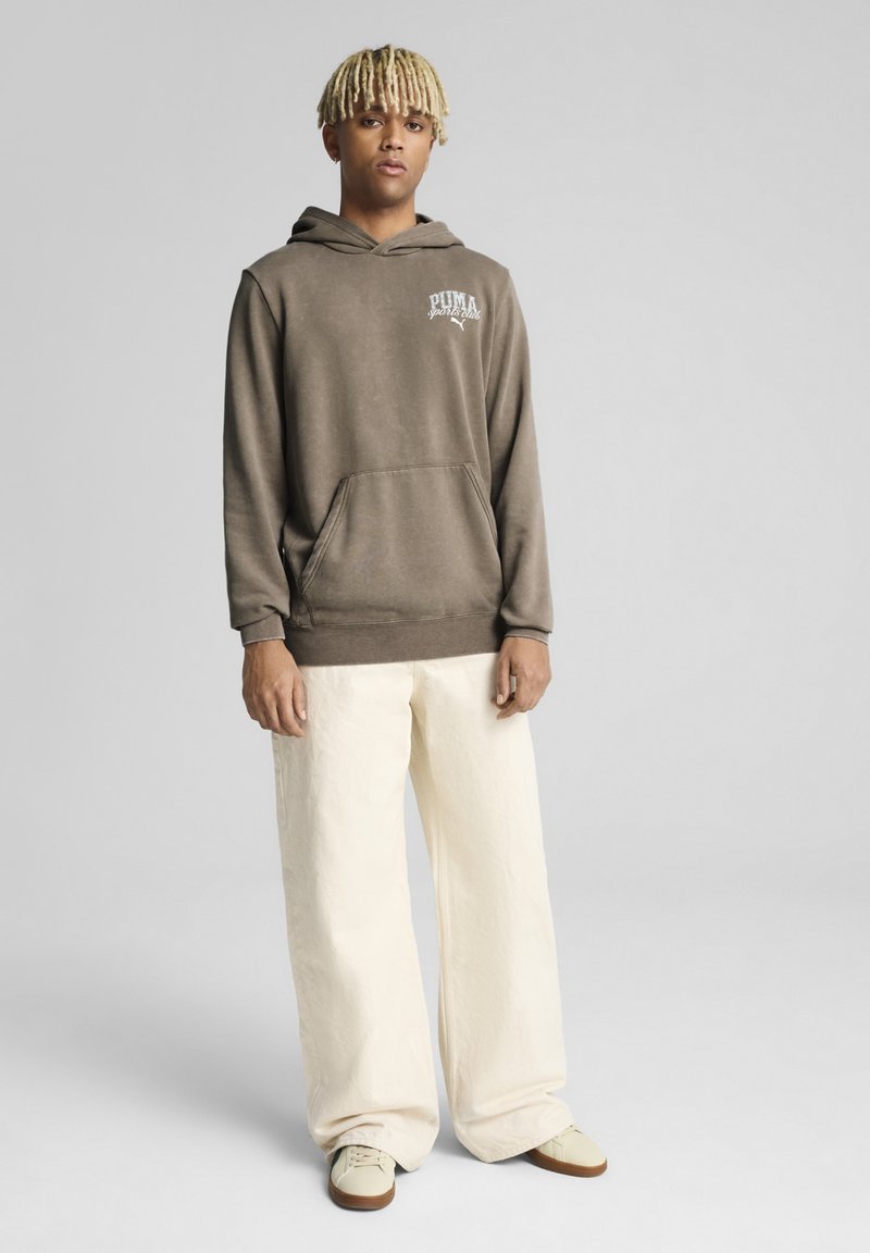 Sudadera con capucha marrón, con bolsillo frontal y logo, combinada con pantalones de pierna ancha color crema. El modelo está de pie sobre un fondo gris.