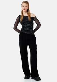 Sort off-shoulder top med gennemsigtige, prikkede lange ærmer og rynket design, parret med højtaljede sorte vide bukser og platform sko.