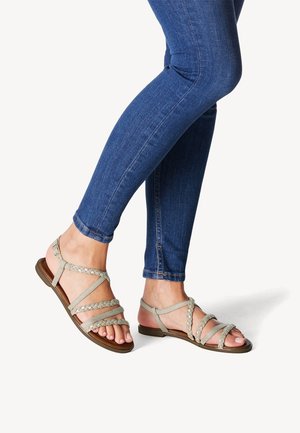 Sandali grigi chiari con cinghie intrecciate e suole piatte, indossati con jeans skinny blu scuro, mostrando le dita dei piedi e il design aperto.