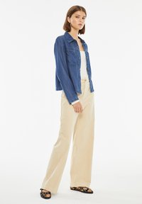 Jeans jakna, svetlo modra, spredaj z gumbi, detajl žepa na prsih; v kombinaciji z bež širokimi hlačami in črnimi čevlji z cvetličnim poudarkom.