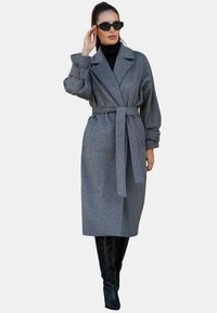 Szary płaszcz trench z mieszanki wełny o kroju kopertowym, z wiązanym w pasie paskiem i przeskalowanymi rękawami. Noszony z czarnym golfem i czarnymi okularami przeciwsłonecznymi.