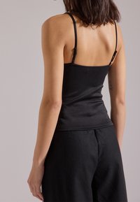 Donna con capelli scuri fino alle spalle, che indossa un top nero con spalline sottili e pantaloni neri, vista da dietro su uno sfondo neutro.