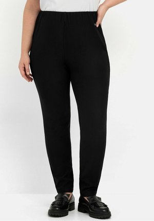 Schwarze gerippte Hose mit elastischem Bund, figurbetonter Passform und glatter Textur, kombiniert mit schwarzen Slippern mit Fransen-Detail.