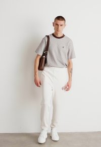 Koszulka w paski z krótkim rękawem z brązowymi akcentami, białe spodnie dresowe z detalem logo, brązowa torba na ramię oraz białe sneakersy.