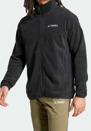 Person wearing a black zip-up fleece jacket and olive green pants, both with white "TERREX" logos, standing against a light background.