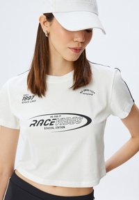 Camiseta blanca recortada con texto y gráficos en negro, incluyendo "RACEVIBES" y "EDICIÓN ESPECIAL". Mangas cortas con acentos en negro.