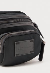 Saco de nylon preto com dois fechos de correr prateados e um patch da logo da Armani Exchange cosido no bolso frontal.