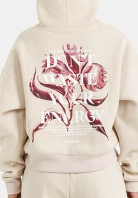 Felpa beige con grafica floreale rosa sul retro e testo. Progettata con una vestibilità rilassata e un bordo ribbed in vita e ai polsini.