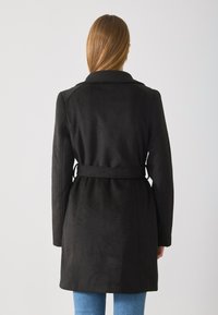 Svart beltefrakk med et mykt, strukturert stoff. Har en notched krage og en tilpasset midje. Rekker til midten av låret, designet med lange ermer.
