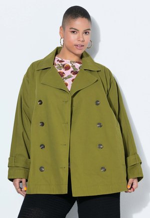 Mujer con una chaqueta de doble botonadura extragrande de color verde oliva sobre una blusa rosa con estampado, posando contra un fondo blanco.