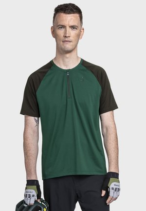 Mann trägt ein grün-schwarzes kurzärmliges Hemd, schwarze Shorts und fingerlose Handschuhe und hält einen passenden Helm vor einem neutralen Hintergrund.