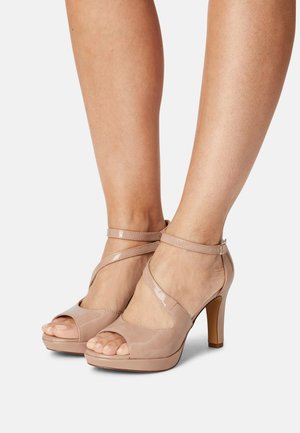 Nude lakierowane sandały na wysokim obcasie z otwartym noskiem, paskiem wokół kostki i platformą. Posiadają krzyżowy design pasków oraz drewniany obcas.