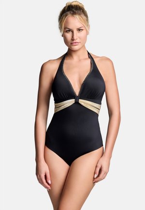 Blondynka z związanymi włosami, ubrana w czarny strój kąpielowy z dekoltem halter i złotym paskiem w talii, stojąca na jednolitym tle.