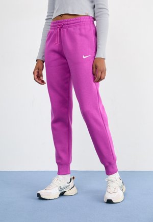 Osoba nosząca jasnofioletowe dresy Nike, białe buty sportowe Nike oraz jasnoszary top z długim rękawem i krótkim fasonie, stojąca na niebieskiej podłodze.