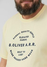 Geel T-shirt met korte mouwen, voorzien van marineblauwe tekst in een cirkelontwerp. De tekst bevat "S. OLIVER A.R.R." en zinnen over kwaliteit en duurzaamheid.