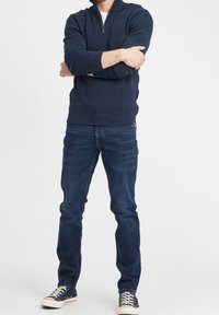 Maglione lavorato a maglia blu navy con colletto a zip, caratterizzato da strisce testurizzate; abbinato a jeans blu scuro e sneakers navy con suole bianche.