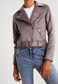 Veste de moto en faux suède gris avec une taille ceinturée, des revers proéminents et des zips argentés ; portée sur une chemise blanche et un jean bleu.