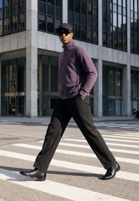 Homme portant une casquette noire, des lunettes de soleil, une veste violette et un pantalon noir traversant un passage piéton en ville avec un immeuble moderne en verre en arrière-plan.