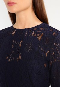 Granatowa koronkowa bluzka z misternymi wzorami kwiatowymi, teksturowanym wzorem z otwartymi detalami oraz okrągłym dekoltem.