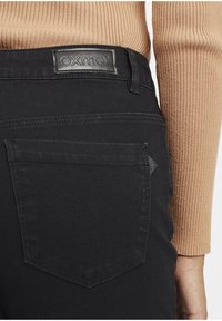 Jean en denim noir avec une surface texturée, comportant une étiquette en cuir avec "OXMO", et une poche arrière avec un accent en forme de diamant.