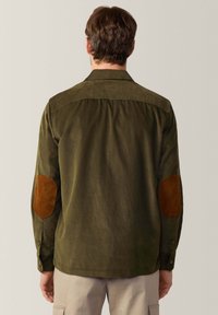 Camicia in velluto a coste verde oliva con toppe in pelle scamosciata beige sui gomiti, polsini bottonati e yoke posteriore dritto. Tessuto testurizzato con una vestibilità rilassata.