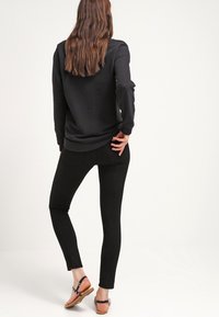 Černá mikina s hladkou texturou, spárovaná s černými skinny džíny a hnědými sandály. Oblečení zdůrazňuje štíhlý siluetu.