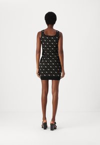 Femme debout de dos vêtue d'une mini robe noire sans manches avec un motif de losanges beige et logos, accompagnée de sandales noires à talons.