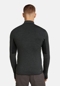 Mężczyzna z krótkimi włosami, ubrany w dopasowaną, ciemnoszaro długą bluzę i brązowe spodnie, stojący tyłem na jednolitym białym tle.