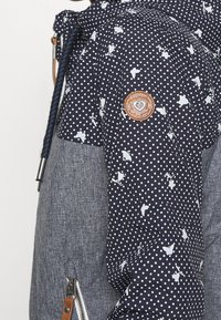 Veste matelassée bleu marine ornée de pois blancs et de motifs floraux, avec une partie inférieure texturée grise et un patch circulaire de la marque sur la manche.