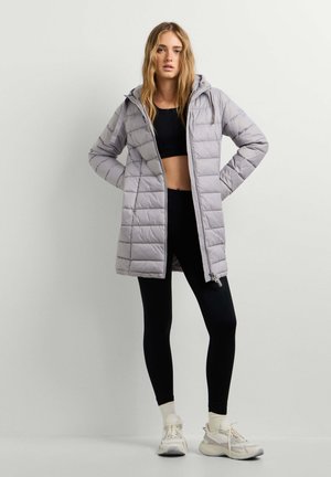 Frau trägt eine hellgraue gesteppte Kapuzenjacke über einem schwarzen Croptop, schwarzen Leggings, weißen Socken und weißen Turnschuhen, steht mit den Händen in der Hüfte.