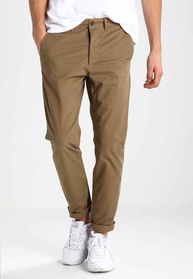 Khaki chino kalhoty s úzkým střihem, hladkou bavlněnou tkaninou, ohrnutými manžetami a předními kapsami, doplněné bílými teniskami.