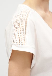 Λευκό πουκάμισο με σύντομο μανίκι, με περίτεχνη κάθετη διακόσμηση από ανοιχτή δαντέλα κατά μήκος του ώμου και ρεβέρ στο μανίκι.