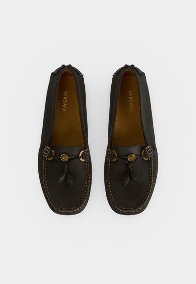 Paire de mocassins en cuir noir avec pampilles, accents dorés, coutures visibles et marque Versace à l'intérieur.