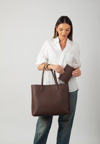 Brown leather tote bag with smooth texture and two handles, accompanied by a matching small wallet, held by a person in a white shirt.