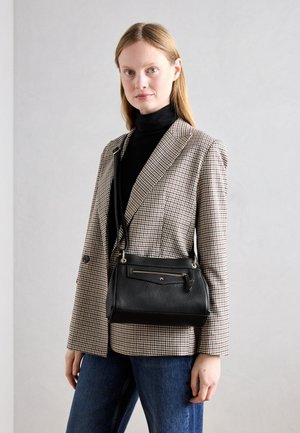 Vrouw draagt een geruite blazer en een zwarte coltrui, met een zwarte leren crossbodytas, staande tegen een effen lichte achtergrond.