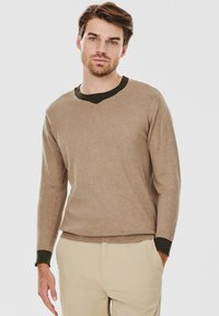 Beżowy sweter wykonany z miękkiego materiału, z dekoltem w kształcie litery V oraz ciemnozielonym prążkowanym kołnierzykiem i mankietami. Długie rękawy i dopasowany krój.