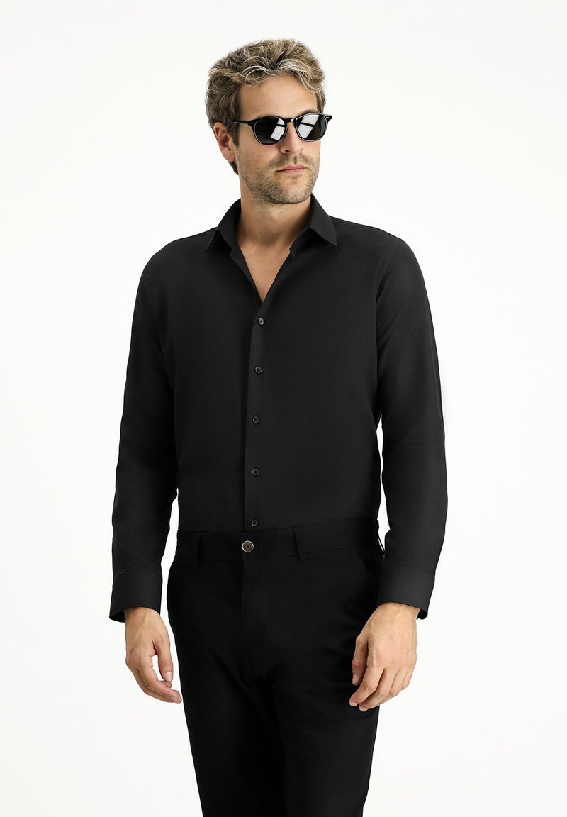Camicetta nera a maniche lunghe con colletto e chiusura a bottoni, abbinata a pantaloni neri. Modello che indossa occhiali da sole scuri.