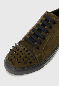 Olivgrön mockasneaker med svart gummisula, med en spetsig tå och sex svarta snören som går genom metallögle. Slät yta hela vägen igenom.