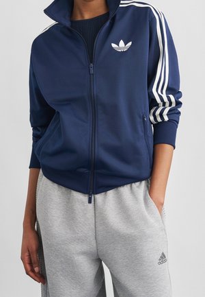 Personne portant une veste de survêtement Adidas bleu marine avec des bandes blanches et un pantalon de survêtement gris Adidas, les mains dans les poches, debout devant un fond uni.