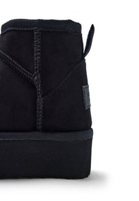 Botín negro de ante con una superficie texturizada, que presenta un detalle de costura prominente y una suela de goma plana. Incluye una lengüeta en la parte trasera.