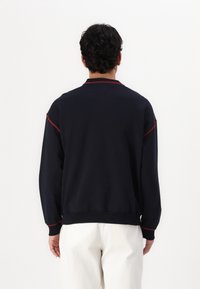 Sudadera navy con acentos rojos, cuello acanalado, mangas largas y un ajuste relajado. Vista desde la parte posterior, mostrando el diseño simple.