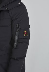 Čierna puffer bunda s kapucňou, na zips a vyšitým logom. Obsahuje textúru tkaniny a elastické manžety pre lepšie prispôsobenie. Nápis SIKSILK na rukáve.