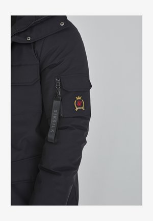 Sort hættetrøje pufferjakke med lynlåslommer og broderet logo. Har stoftekstur og elastiske manchetter for bedre pasform. SIKSILK-mærke på ærmet.