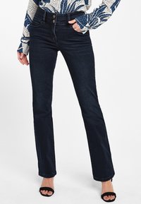 Kobieta w ciemnoniebieskich jeansach bootcut, wzorzystej bluzce i czarnych sandałach na obcasie, stojąca z jedną ręką w kieszeni.