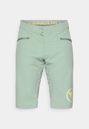 Lys grønne sykkelshorts med en glatt tekstur, to glidelåslommer, perforeringer på sidene og en kontrasterende gul logo.