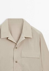 Camicia beige a maniche corte realizzata in tessuto morbido, con colletto classico, taschino e chiusura con bottoni.