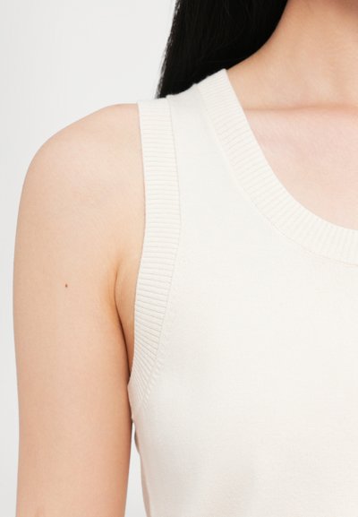 Lys beige ermeløs topp med ribbestrikket utringning og armhuler, laget av et glatt, elastisk materiale med en tettsittende silhuett.