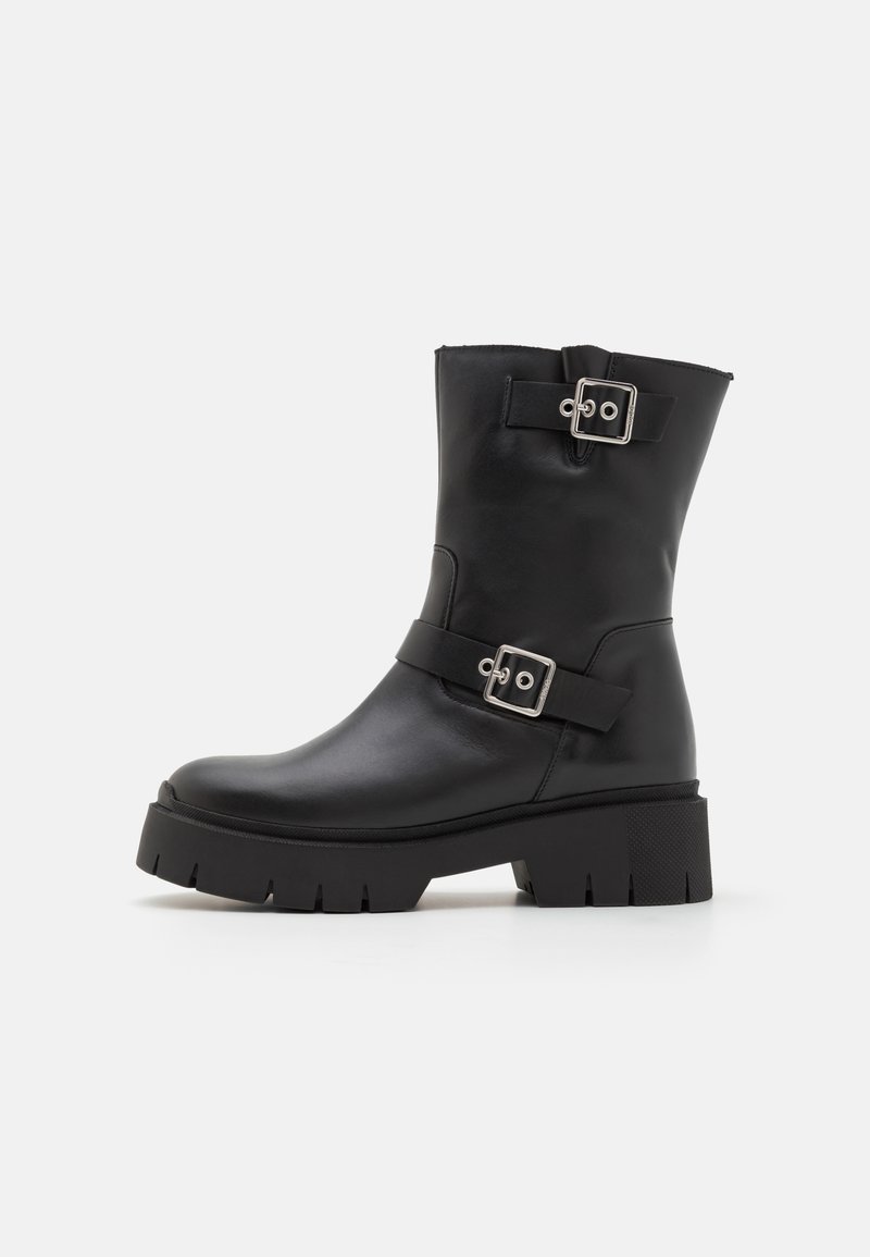 Botas de tornozelo em pele preta com sola robusta, apresentando duas correias com fivelas prateadas. Acabamento suave e textura robusta na sola.