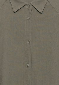 Napitetty oliivinvihreä paita, jossa on kärjistetty kaulus ja näkyvä ommel nappilistan varressa.